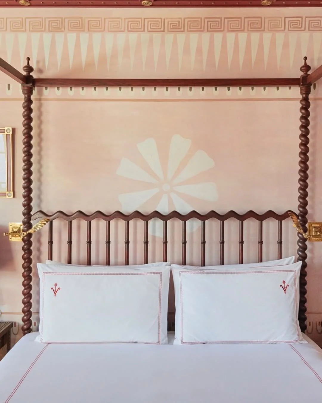 A neatly made bed with white pillows and sheets, featuring red embroidered details. The wooden four-poster bed has twisted posts and gold accents, with a pink wall behind it. The wall has a decorative pattern and an abstract flower design.