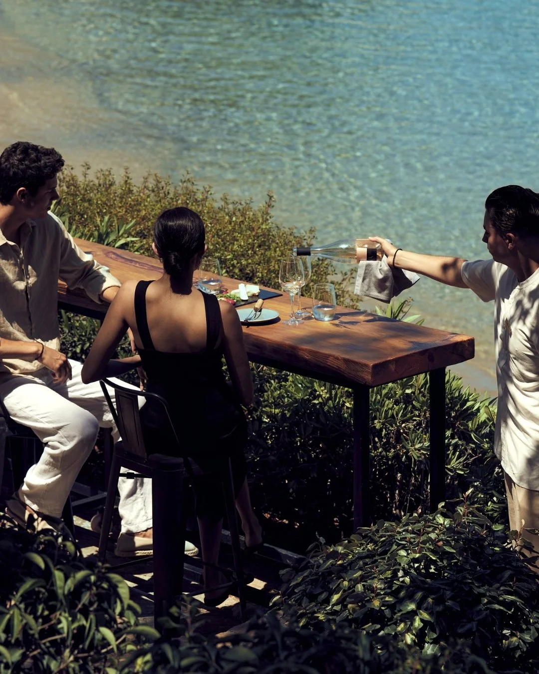 Three people having a meal outdoors by the water. Two men sitting next to each other, and a woman with her back to the camera. A person on the right is pouring a drink.