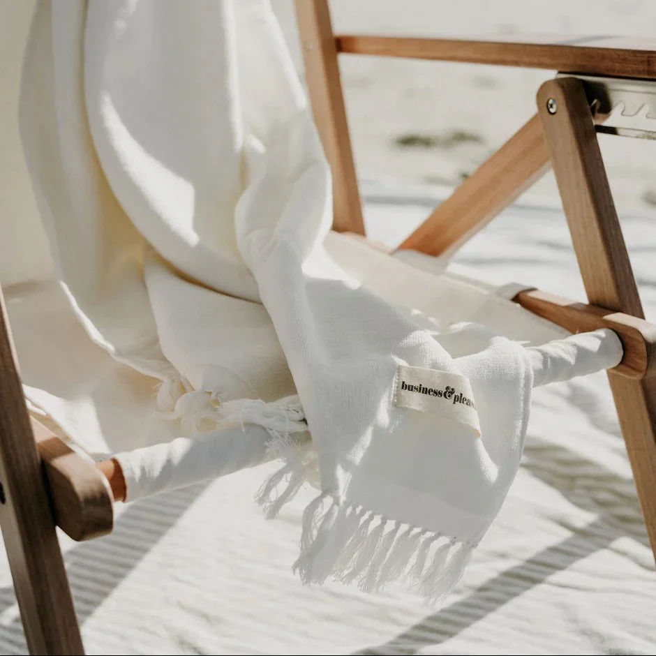 White cloth with the words ''business & pleasure'' on a tag, draped over a wooden folding chair on a bright, outdoor setting.