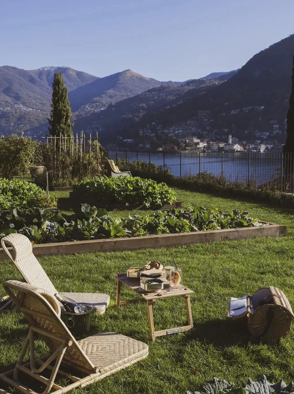 A garden patio with two lounge chairs, a small table with food and drinks, and a view of a lake and mountains in the background.