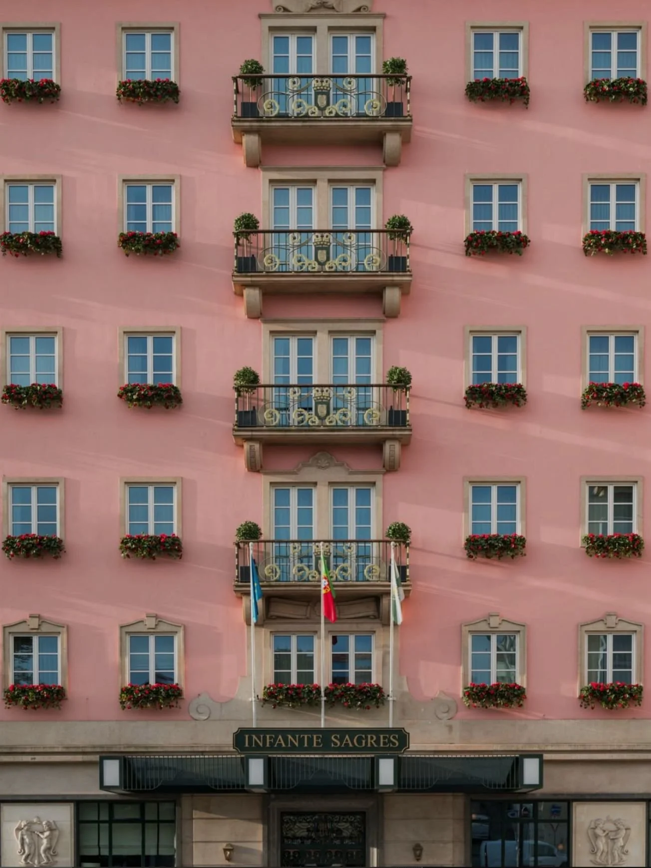 A pink building with multiple windows, decorated with flower boxes filled with red flowers, and several balconies with potted plants. There are flags hanging in front of the building and a sign that reads 'Infante Sagres' above the entrance.