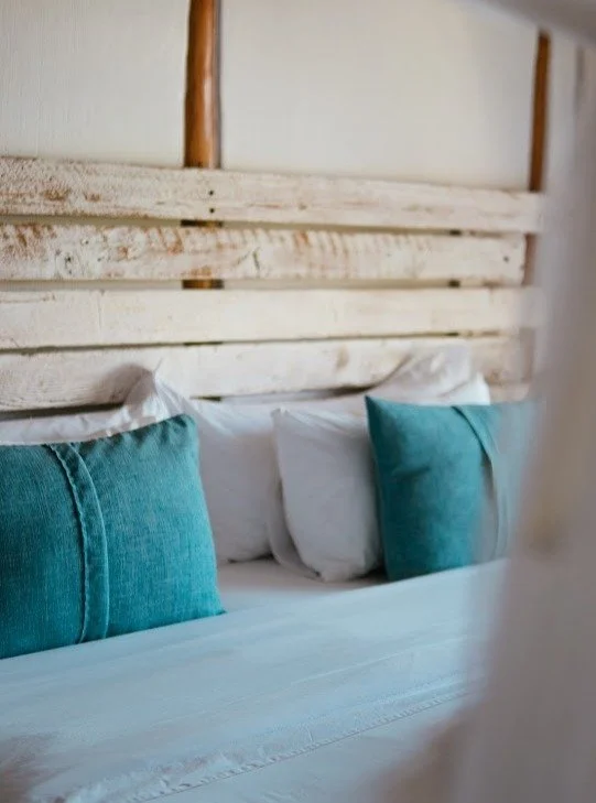 Close-up of a bed with white sheets, two white pillows, and two teal throw pillows, with a rustic wooden headboard in the background.