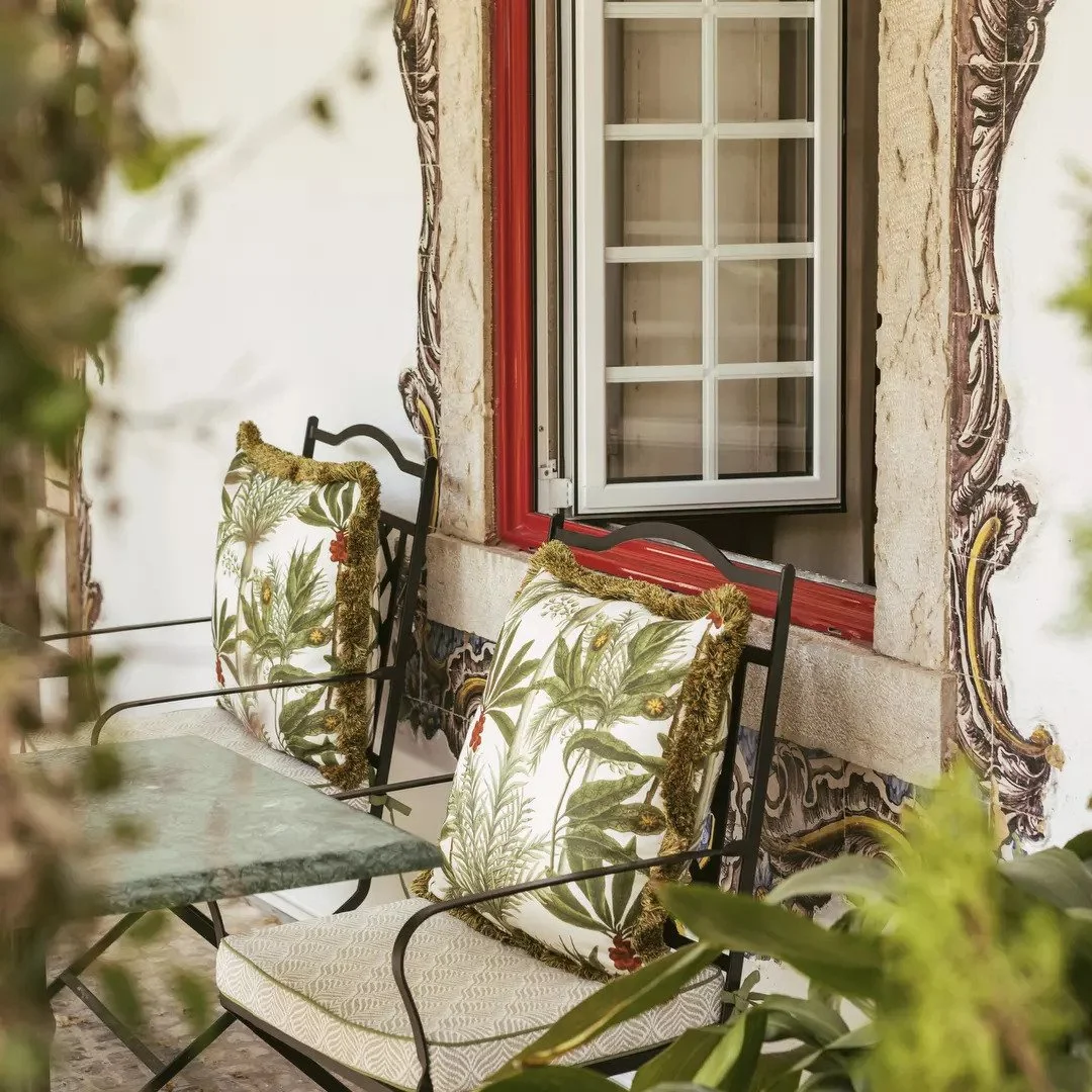 A cozy outdoor patio with two black metal chairs with floral cushions featuring green plants and flowers, and a small glass table, next to a stone and stucco wall with decorative painting details, and an off-white window with a red frame.