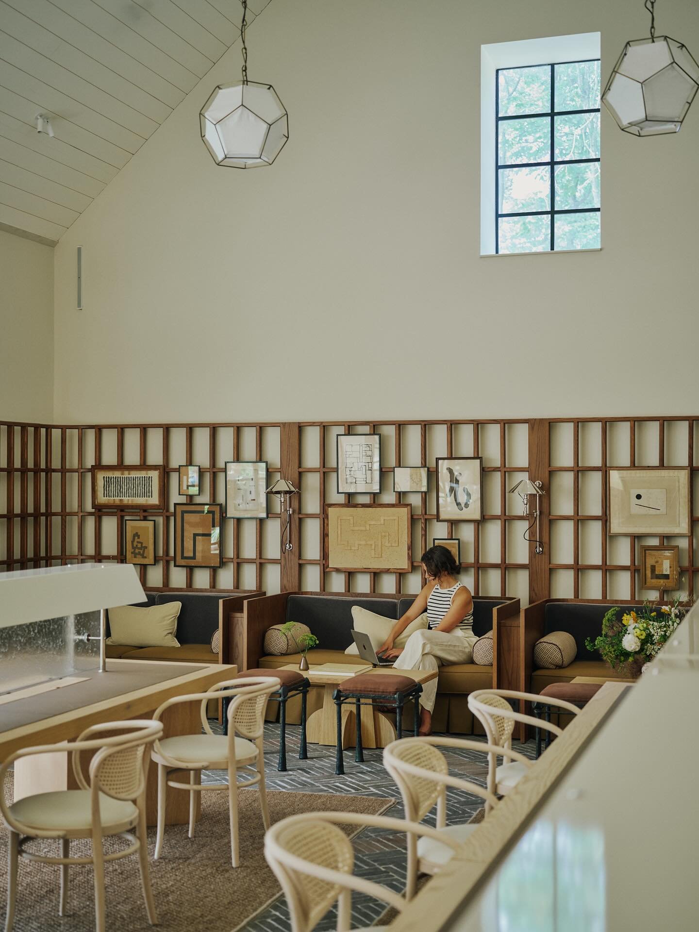 A woman sitting on a sofa in a modern, well-lit coffee shop or lounge area, working on her laptop. The wall behind her is decorated with various framed artwork, and there is a window allowing natural light to illuminate the space.
