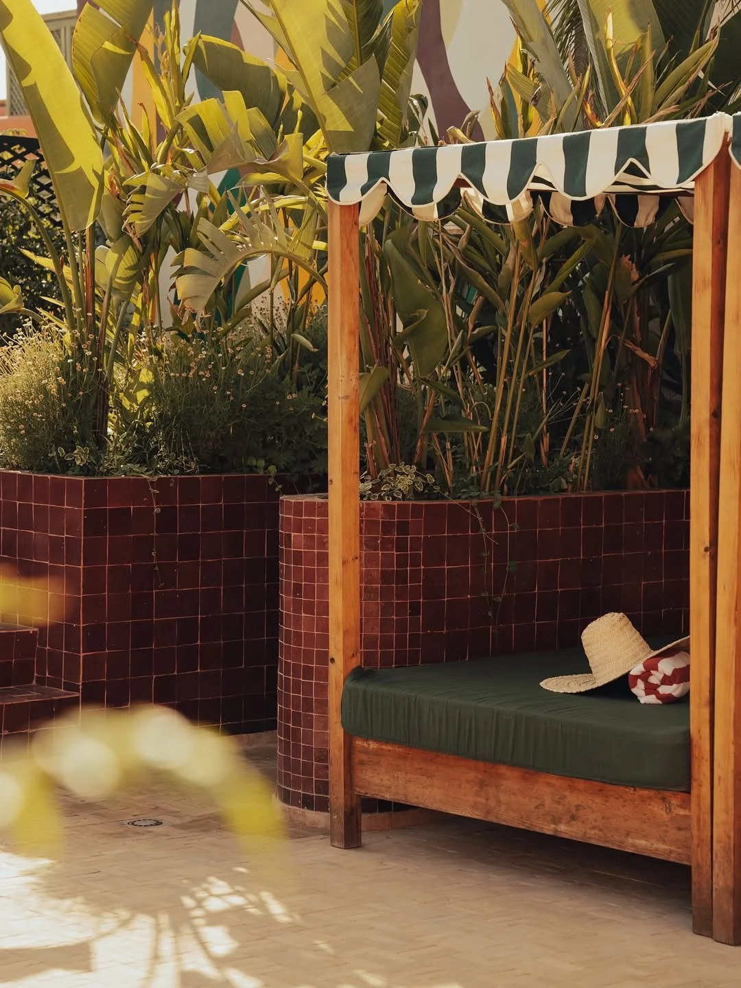 Outdoor seating area with a green cushioned bench under a small striped canopy, a straw hat, and a rolled-up towel, with lush tropical plants in the background.