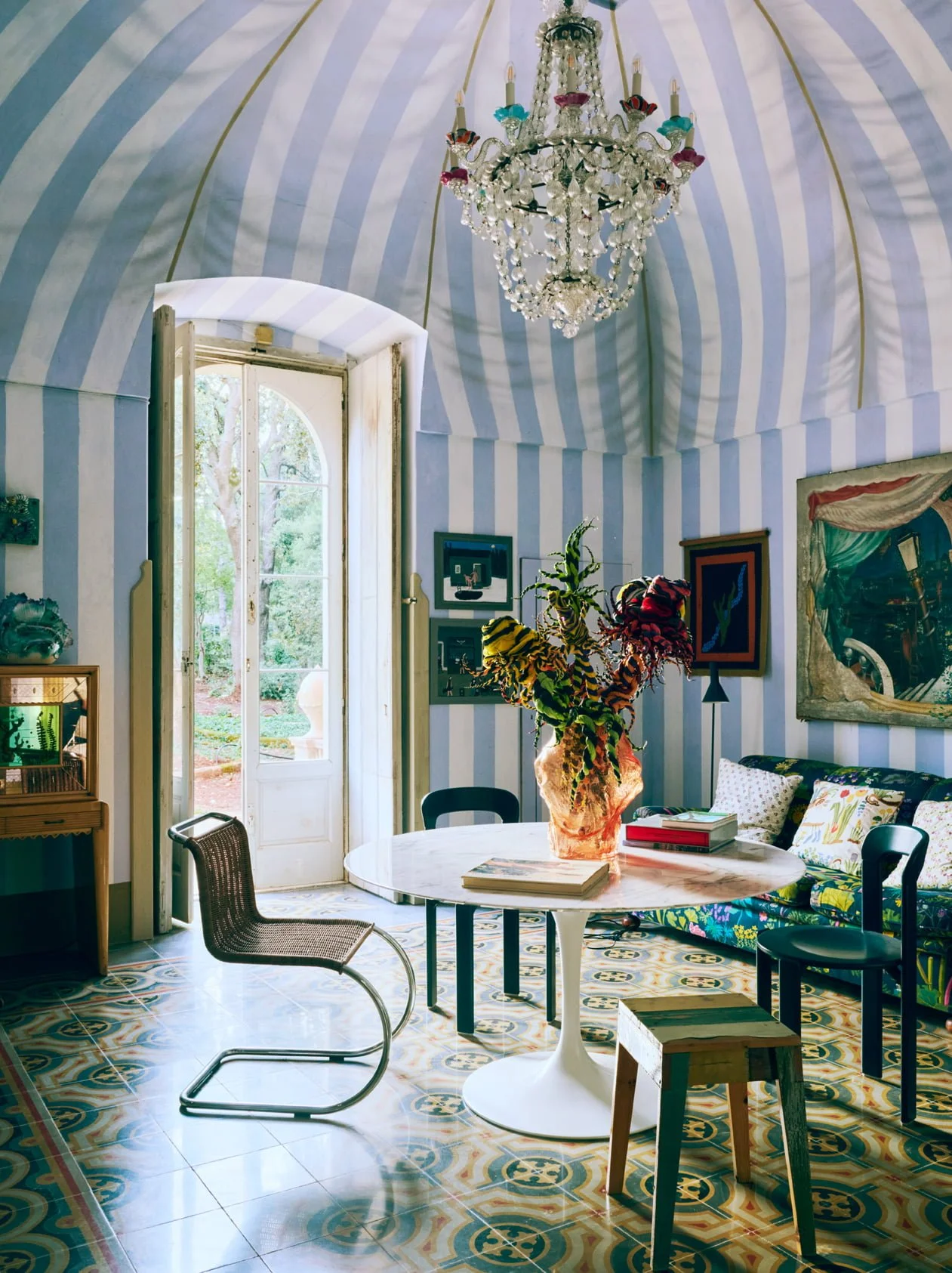 Bright living room with blue and white striped walls, ornate chandelier, open door leading outside, modern and vintage furniture, colorful artwork, and a large floral arrangement on a round table.