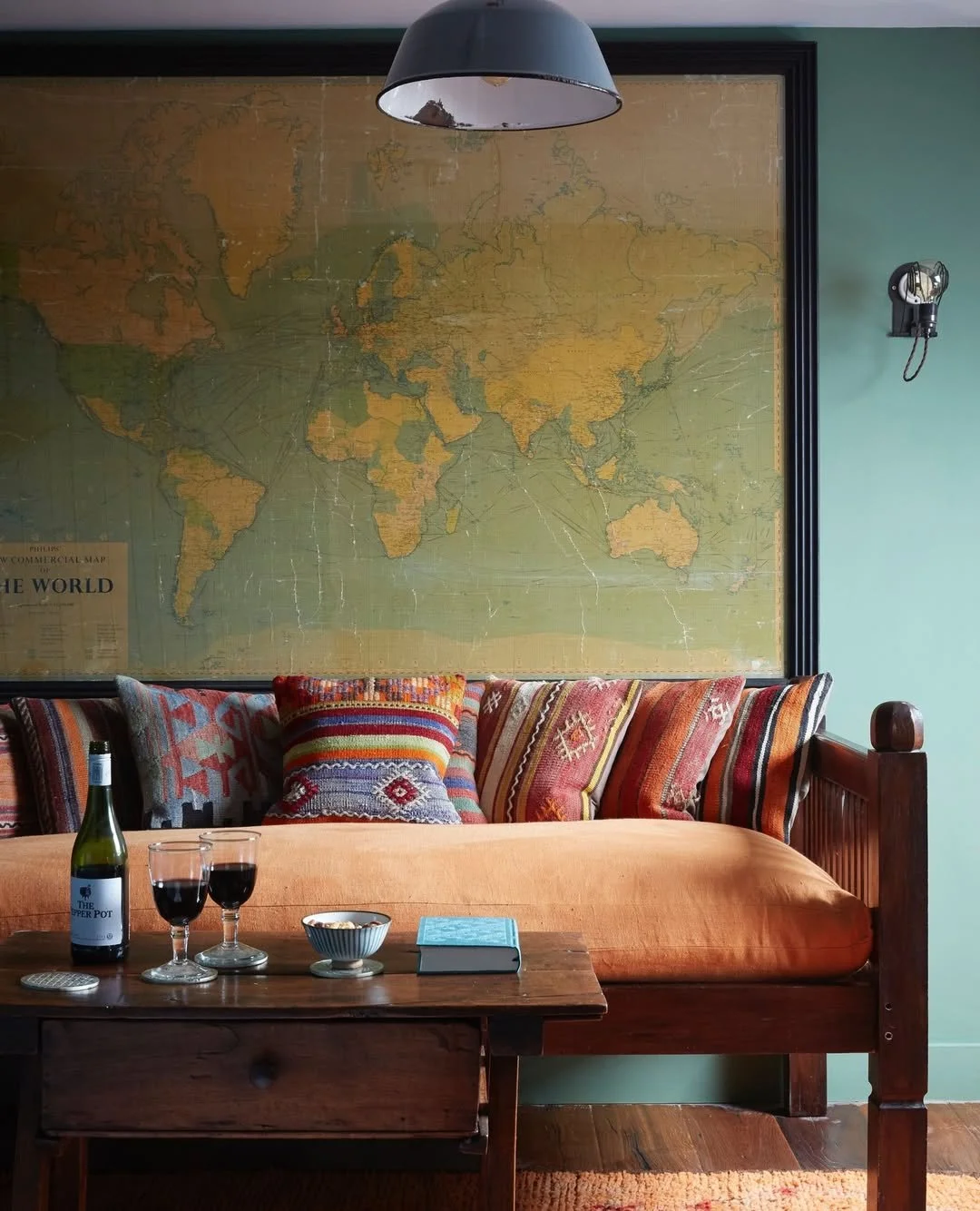 A cozy living room with a large vintage world map on the wall, colorful patterned cushions on a wooden bench, a wooden table with a bottle of wine, two glasses of red wine, a bowl of snacks, and a closed book. The room has a green wall, a black hanging light, and a retro wall phone.