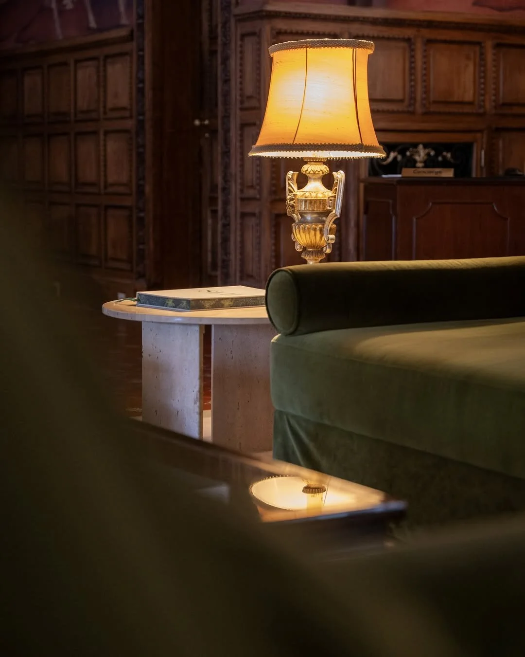 Elegant living room with a classic table lamp with a gold base and warm yellow shade, green velvet sofa, and a round marble side table with a closed book on it, wood-paneled walls in the background.