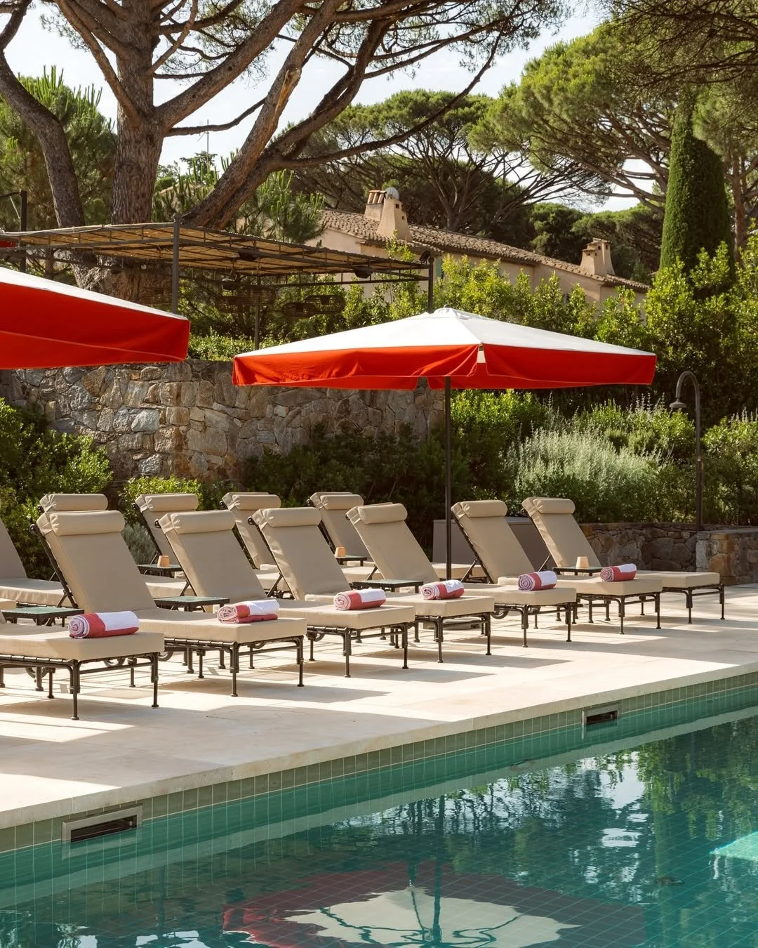 Poolside view with six lounge chairs, each with a rolled towel, under red umbrellas, surrounded by greenery and large trees.