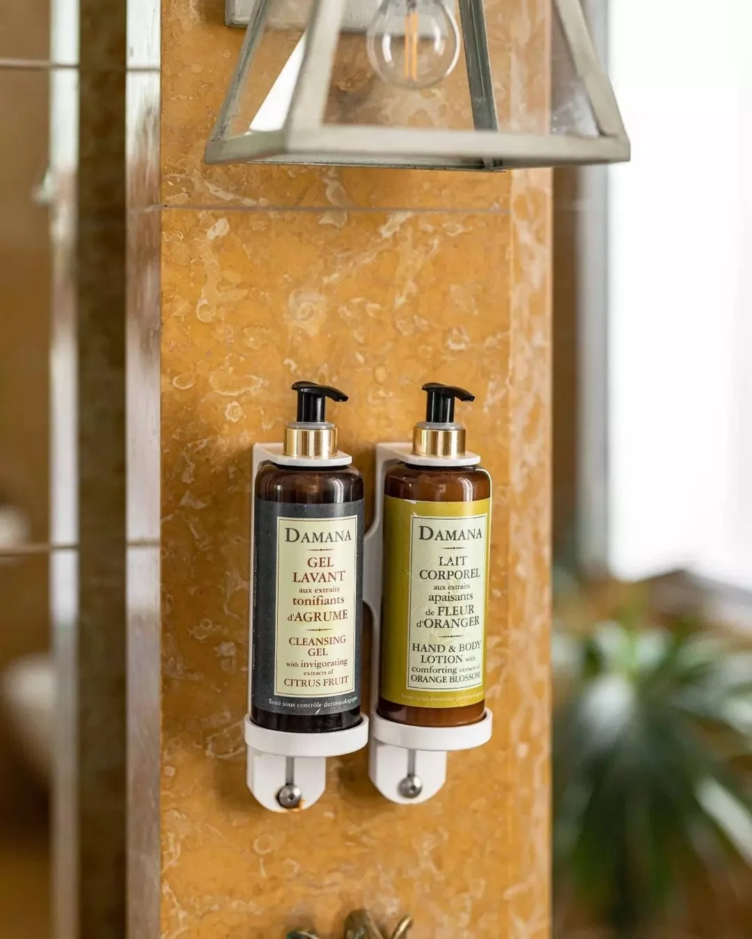 Two brown bottles of Damana toiletries mounted on a peach-colored tiled wall, one labeled 'Cleansing Gel with citrus fruit extract' and the other labeled 'Hand & Body Lotion with orange blossom'.
