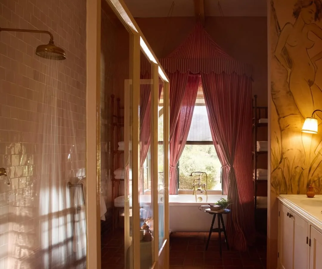 A cozy bathroom with a bathtub in front of a large window with pink and red curtains, a small chair with a plant on it beside the tub, and a shower area with beige tiles.