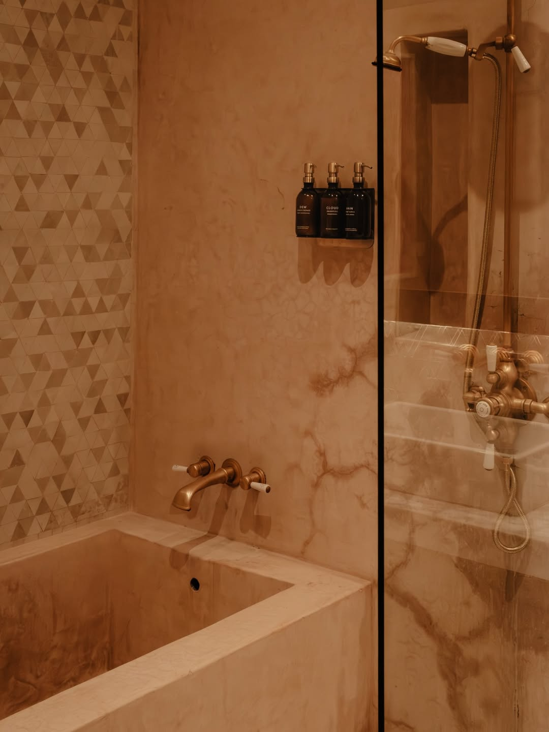 Bathroom with marble walls, a built-in bathtub, a brass faucet, and a glass shower partition with showerhead and control knobs. Bottles of toiletries are mounted on the wall.