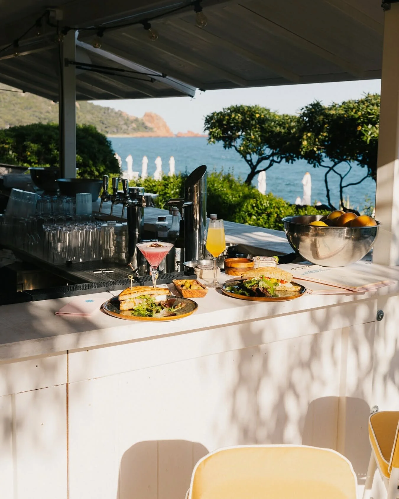 Beachside bar with drinks and sandwiches on a counter overlooking the ocean with trees and parasols in the background.