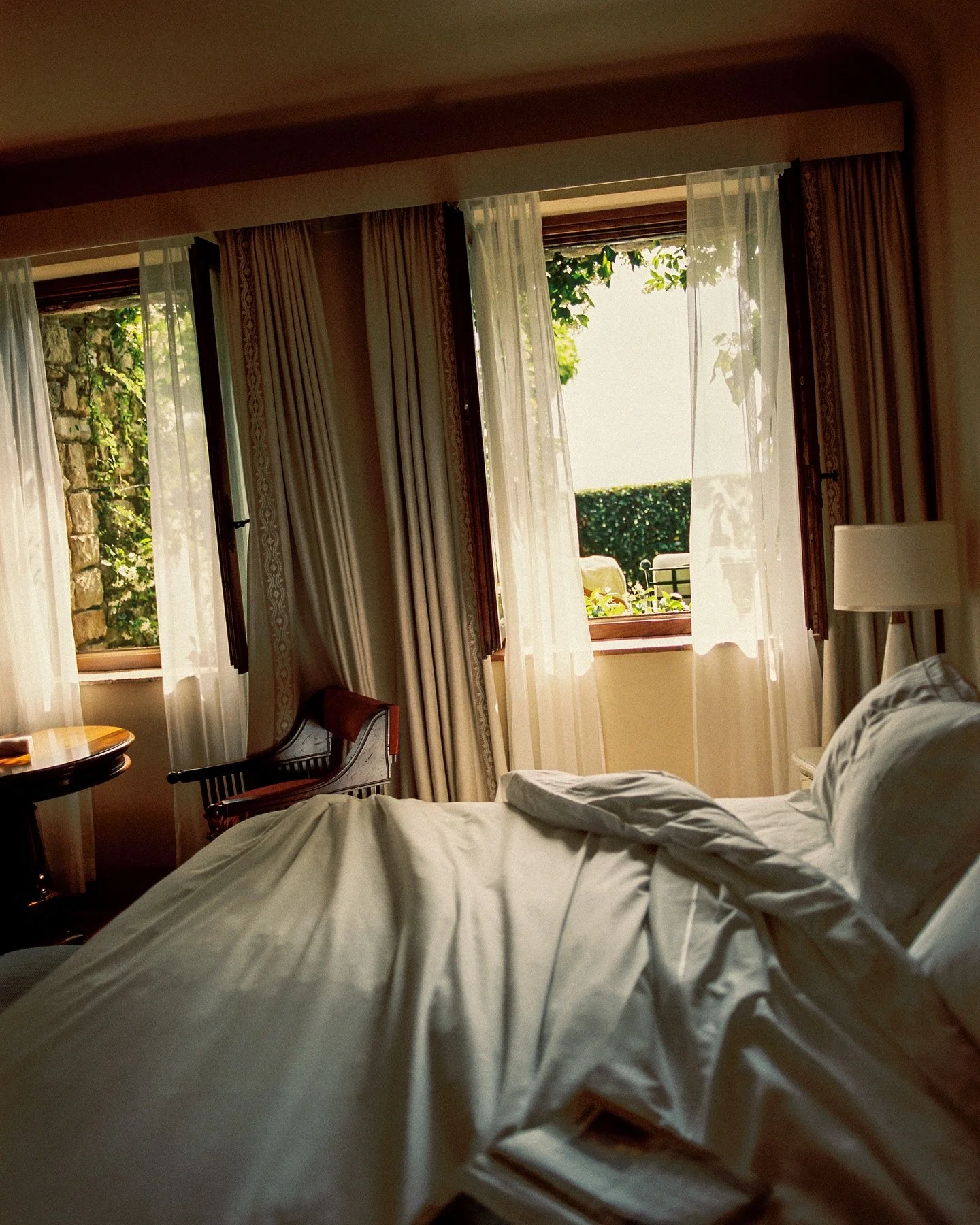 A bedroom with an unmade bed, open windows with sheer curtains, and greenery outside.