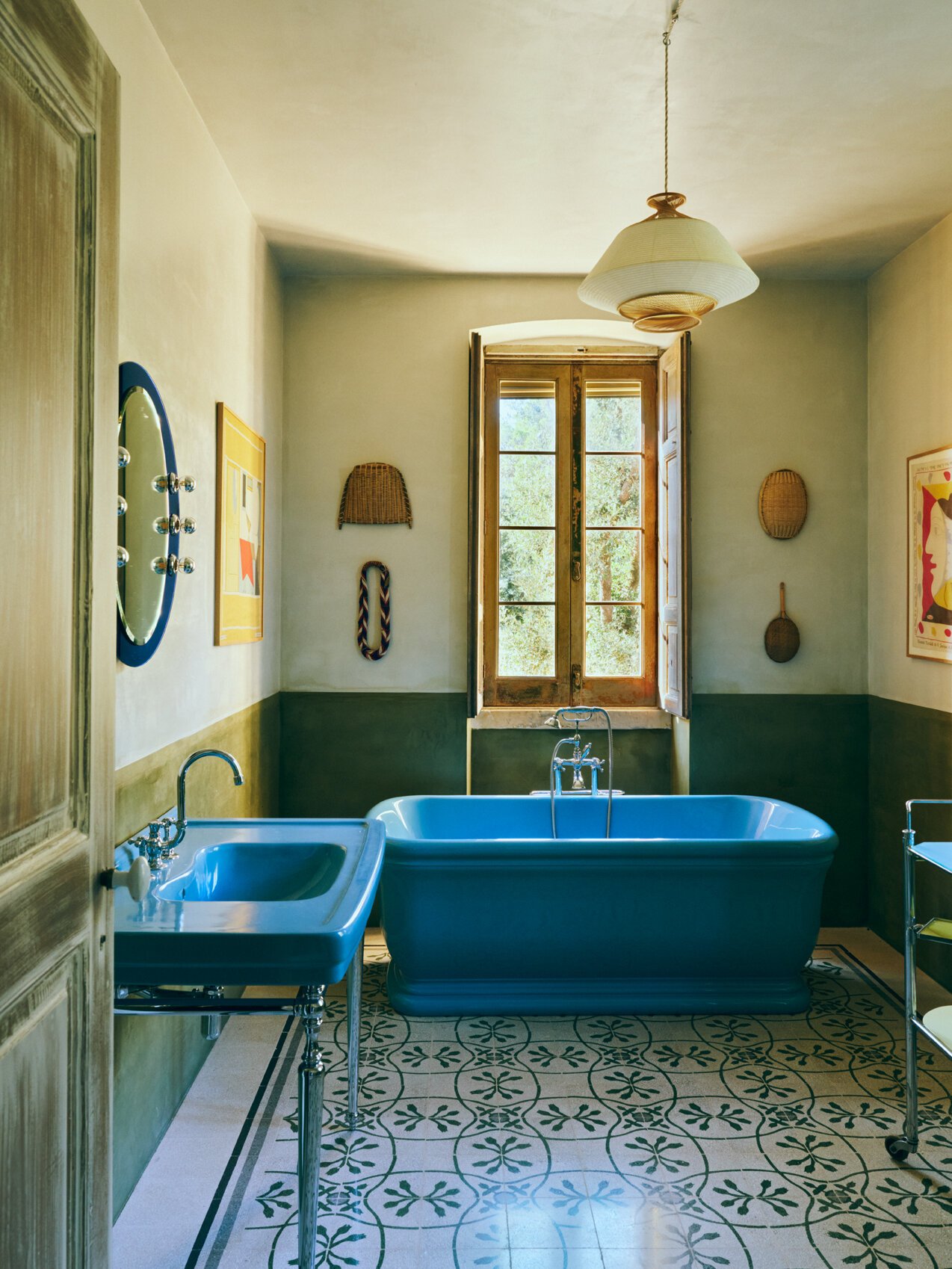A vintage-style bathroom with a blue bathtub, a matching washbasin, a window with wooden shutters, and patterned tile flooring.