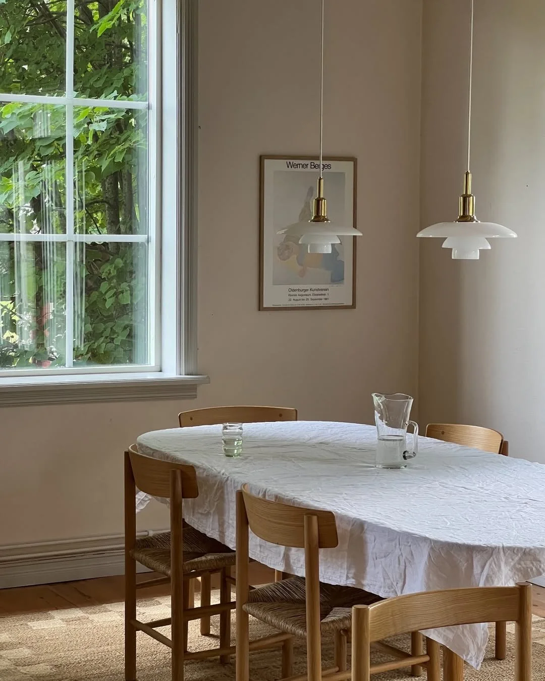 Dining room with a wooden table covered with a white tablecloth, four wooden chairs, a glass water pitcher, a candle, and a framed art poster on the wall. There are two white pendant lights hanging above the table and a window with greenery outside.