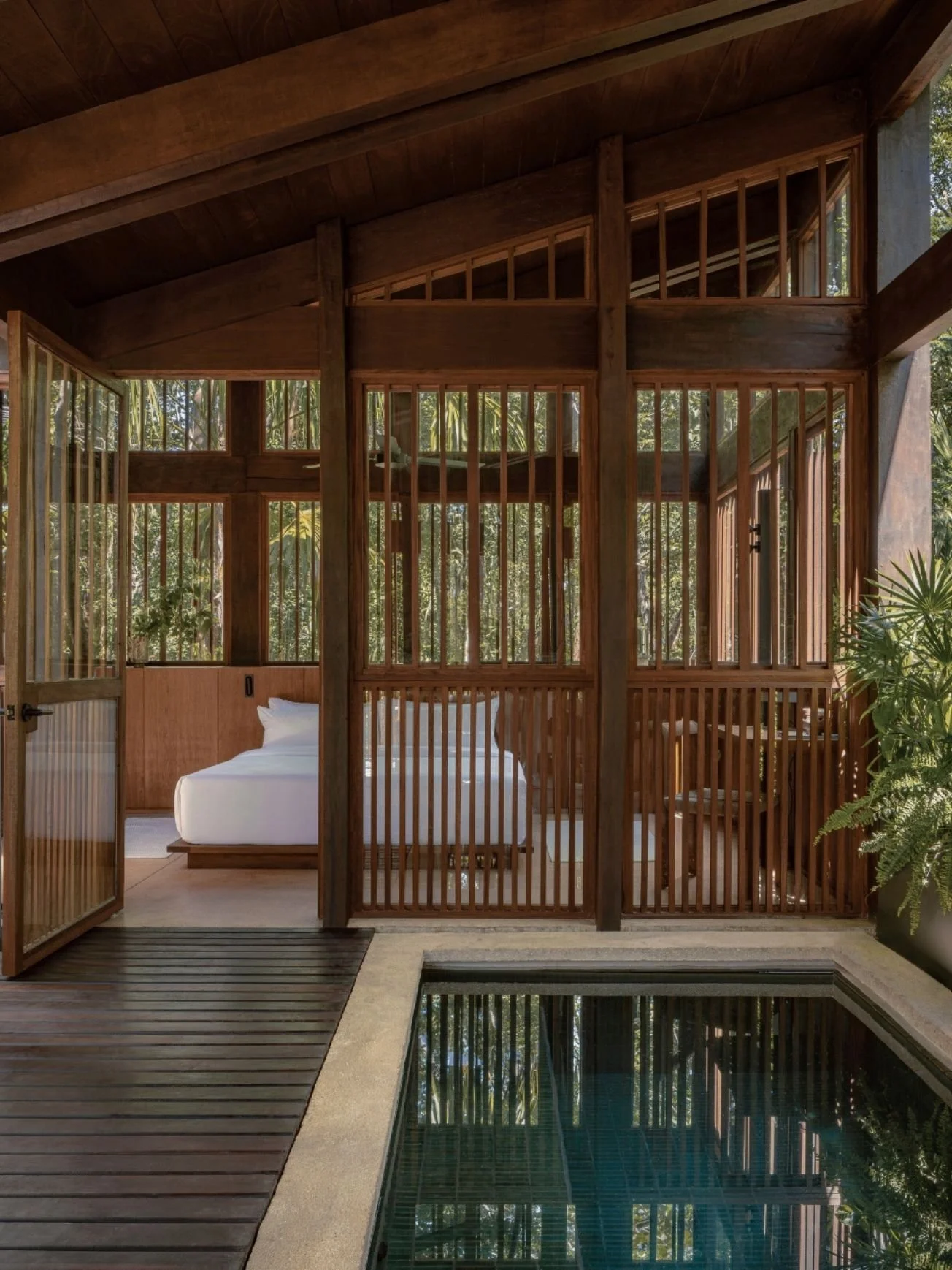 View from outside a wooden structure showing a bedroom with a white bed, glass walls, greenery outside, and a small pool in the foreground.