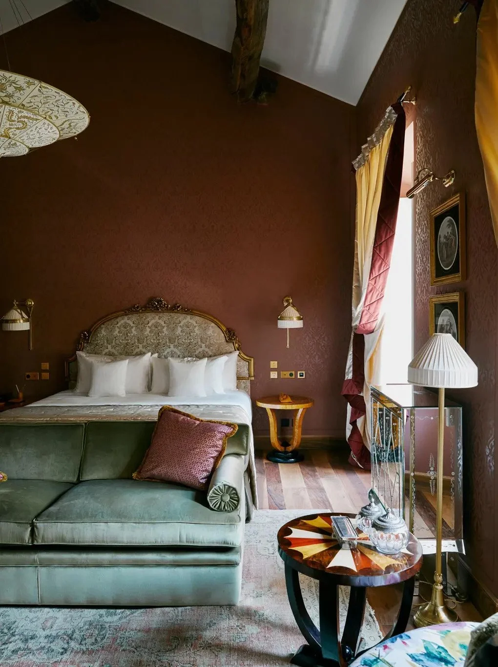 Luxurious bedroom with ornate headboard, green velvet sofa, and warm-colored walls decorated with framed artwork and patterned curtains.