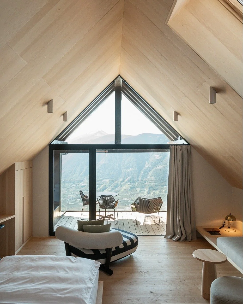 A bedroom with a sloped wooden ceiling, large triangular glass window, and balcony with mountain views. The room has modern furniture, including a black and white striped lounge chair, a bed, a side table, and a desk lamp. Two chairs are on the balcony.