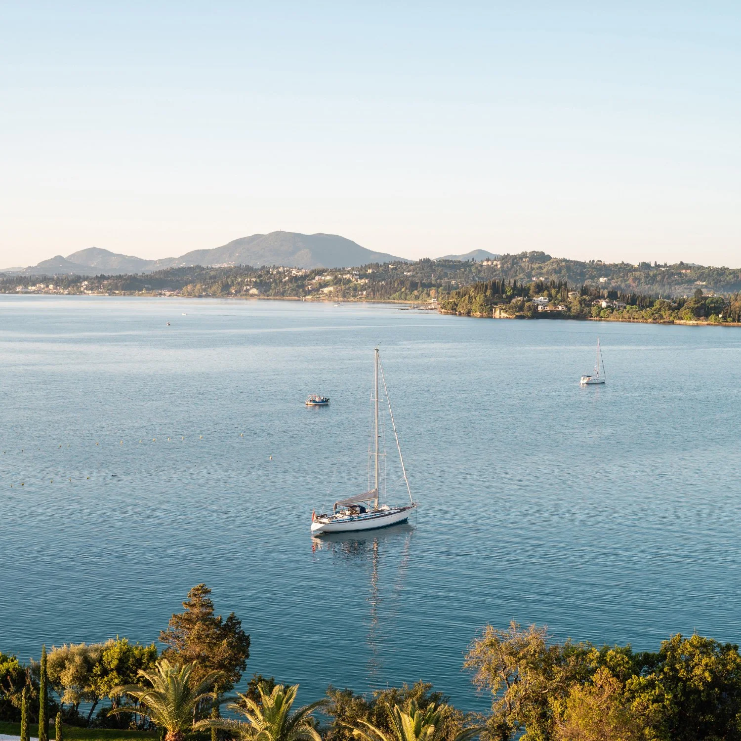 A calm body of water with a sailboat and a motorboat, surrounded by lush green trees in the foreground and a distant hillside with houses and mountains in the background.