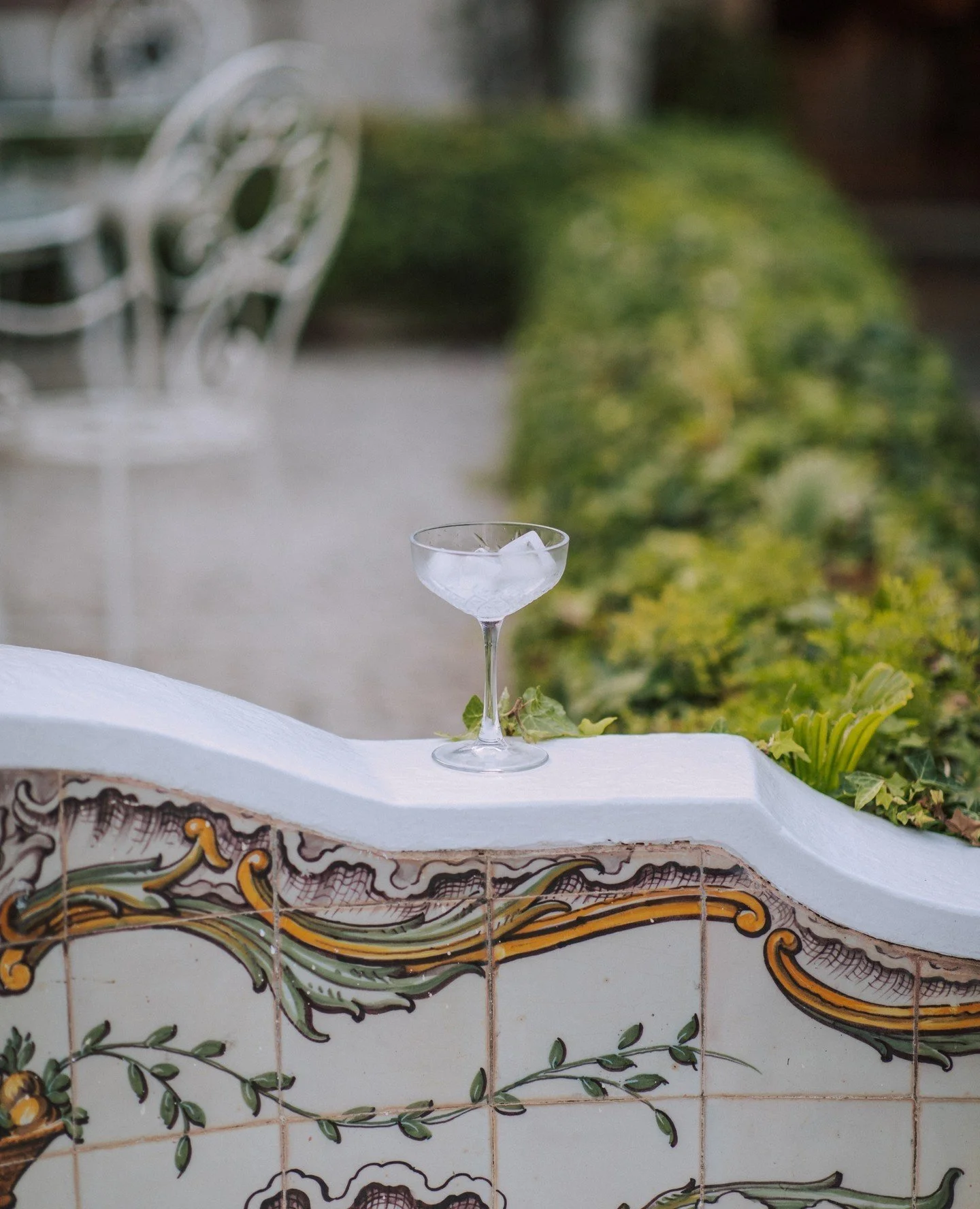 A drinking glass with ice on a white surface, outdoors with greenery and ornate decorated tile wall in the background.