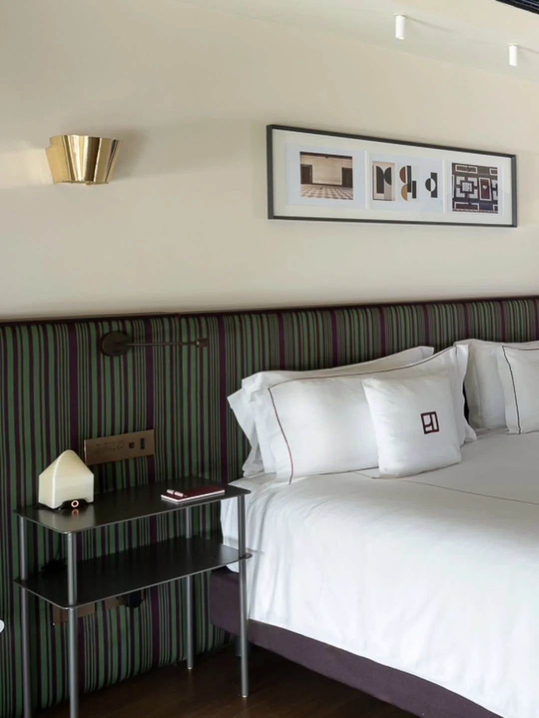 Interior of a hotel room with a bed with white linens and pillows, a striped headboard, a bedside table with a lamp and a small device, a framed abstract art piece above the bed, a wall-mounted reading light, and modern decor elements.