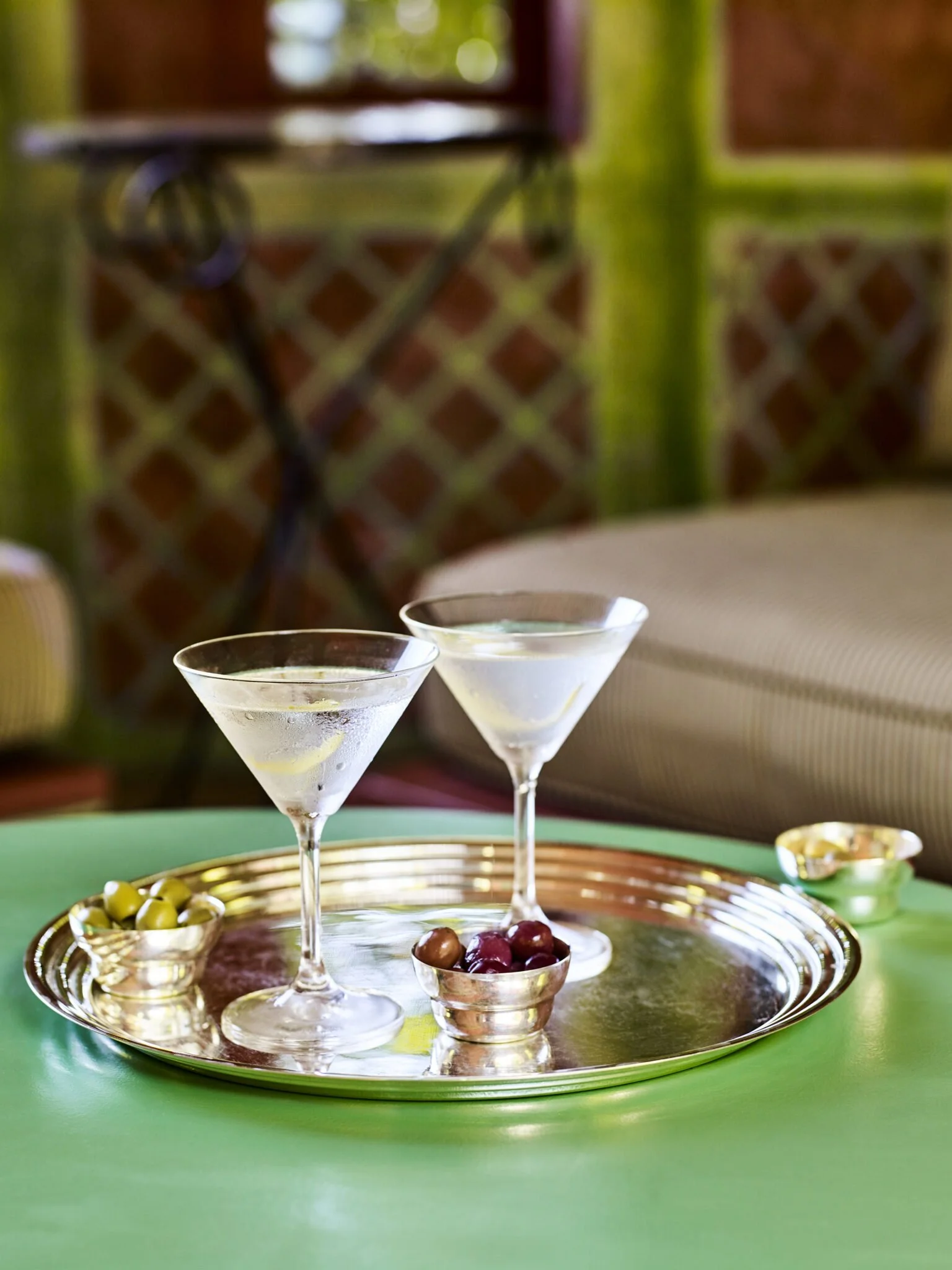 Two martini glasses filled with a clear cocktail and lemon slices, placed on a decorative tray surrounded by bowls of olives and cherries, with a blurred background of a cozy indoor space.