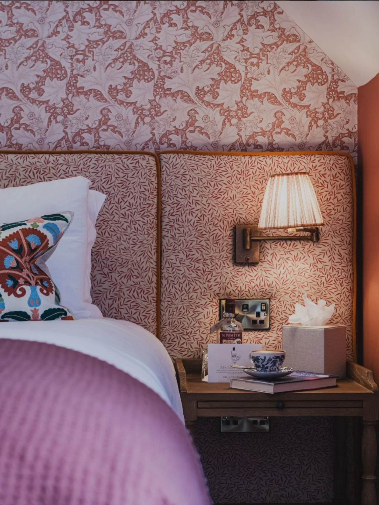A cozy hotel room with a bed that has white bedding, a decorative pillow with colorful embroidery, and a pink textured headboard. There is a bedside table with a lamp, tissue box, a bottle of water, and a mug on top. The wall behind the bed features floral and leaf-patterned wallpaper in shades of pink and white.