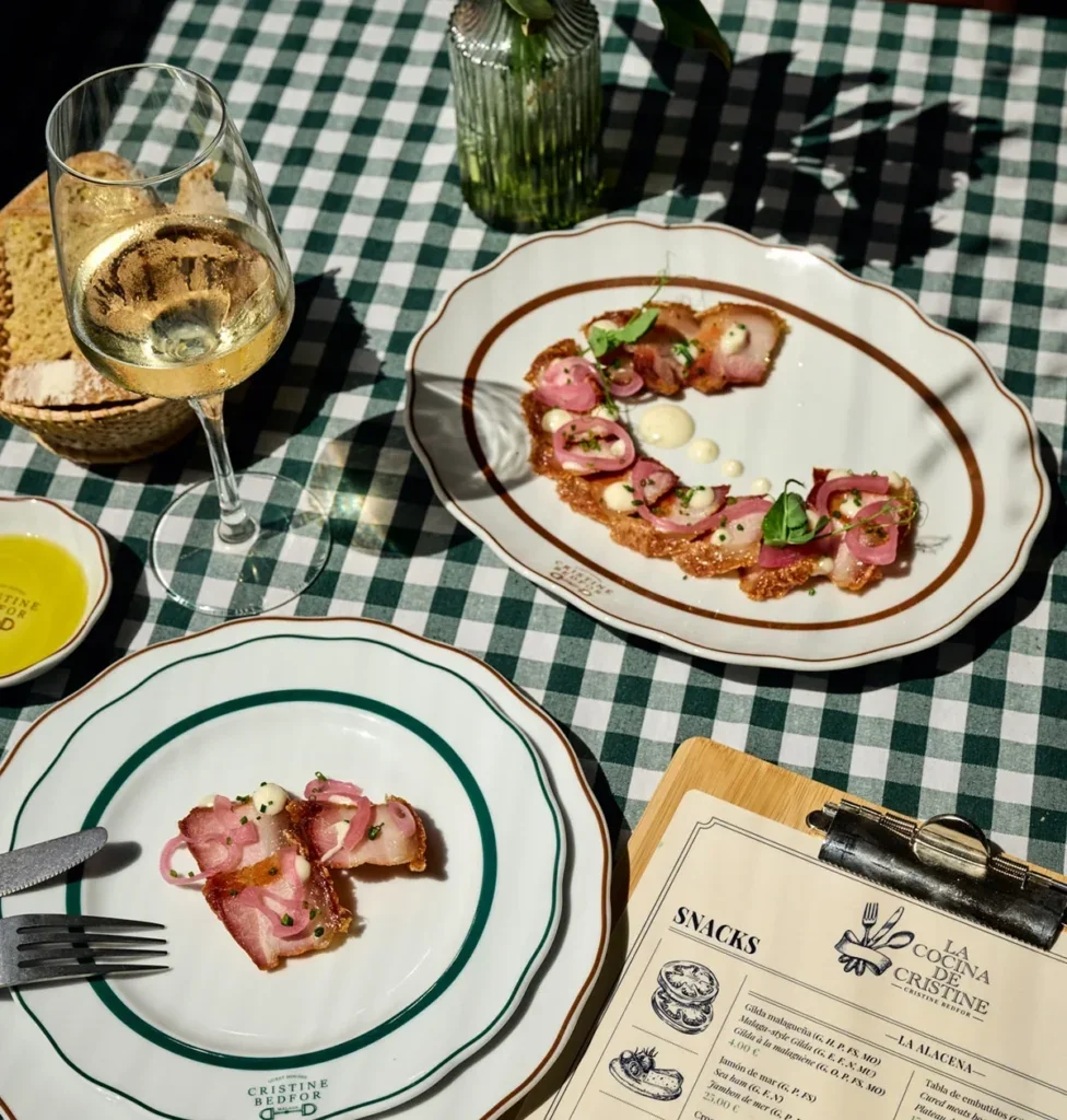 Plate of sliced meat, garnished with herbs and sauce, on a checkered tablecloth, with a glass of white wine and a basket of bread nearby.