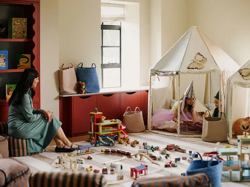 A woman sitting on a couch and two children in a play tent with toys and building blocks scattered on the floor.