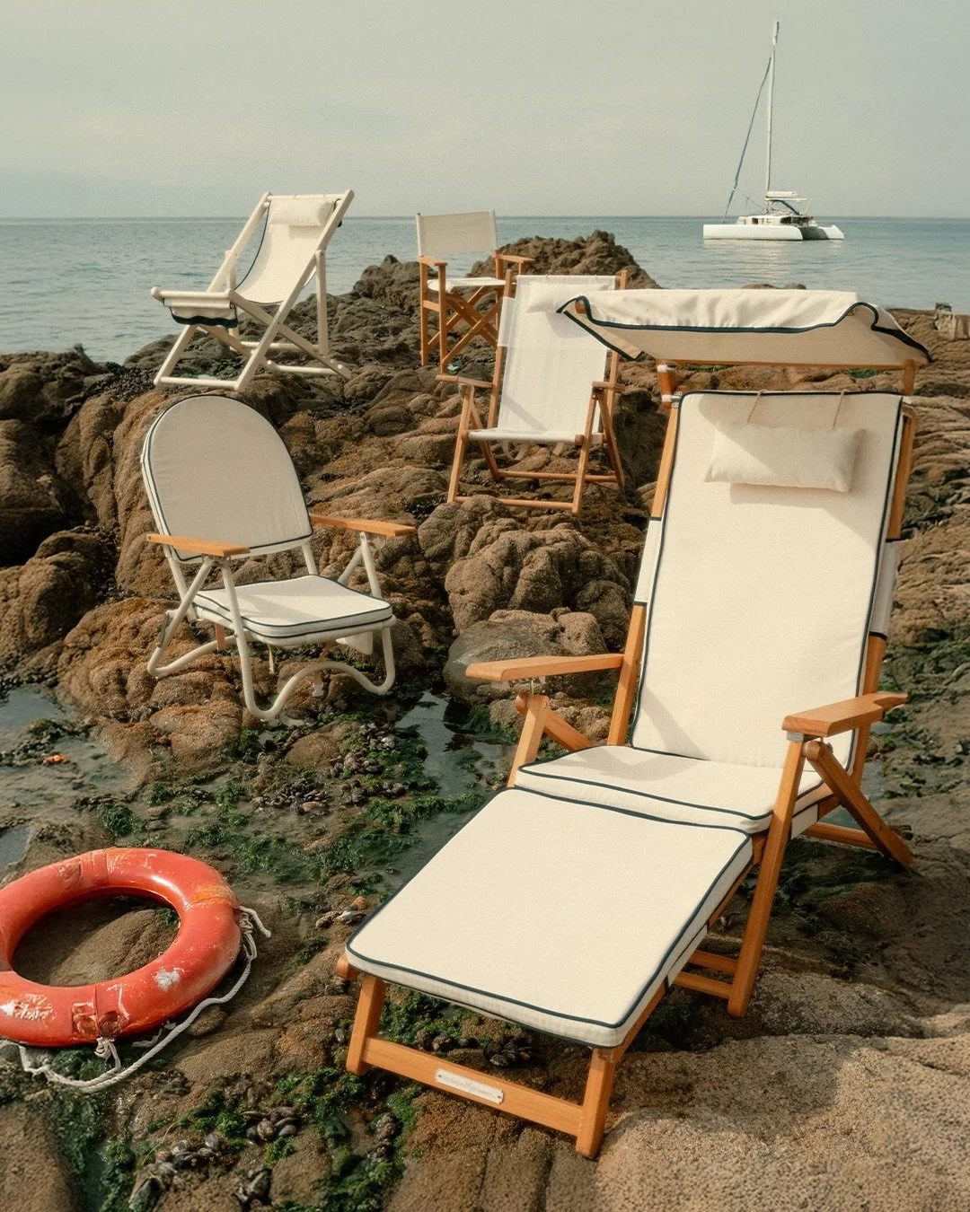 Beach chairs and a life preserver on rocks by the ocean with a sailboat in the background.