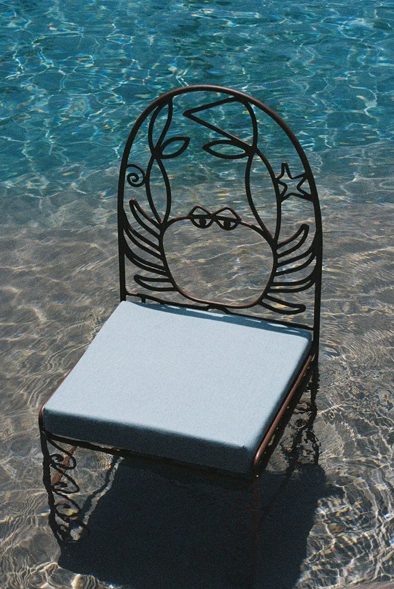 A black metal chair with a decorative face design on the backrest, placed in shallow water with a white cushion on the seat.
