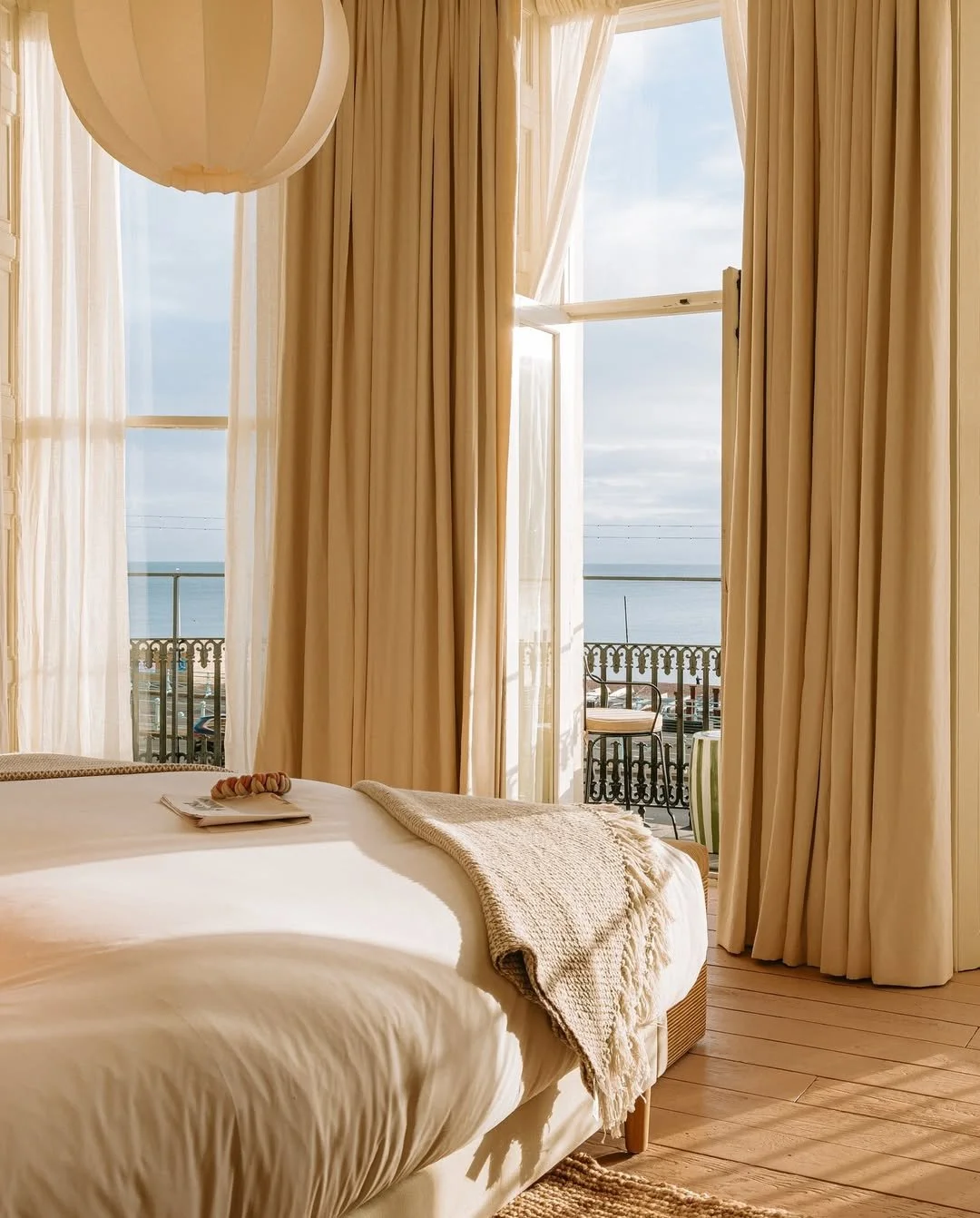 A cozy bedroom with cream-colored curtains and bedding, overlooking a balcony with a view of the ocean.