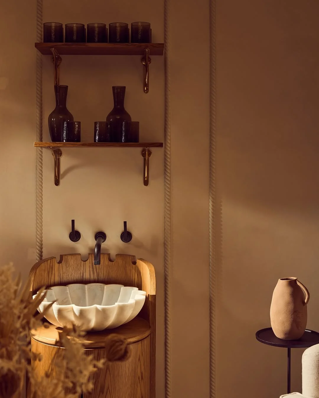 Decorative interior scene with a wooden sink console, a scalloped white vessel sink, dark brown vases on wooden shelves, and a beige ceramic vase on a small black table.