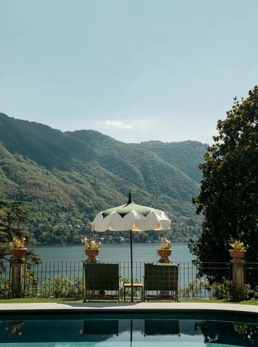 A scenic lakeside view with two chairs and a large patio umbrella overlooking mountains, water, and a railing with potted plants.