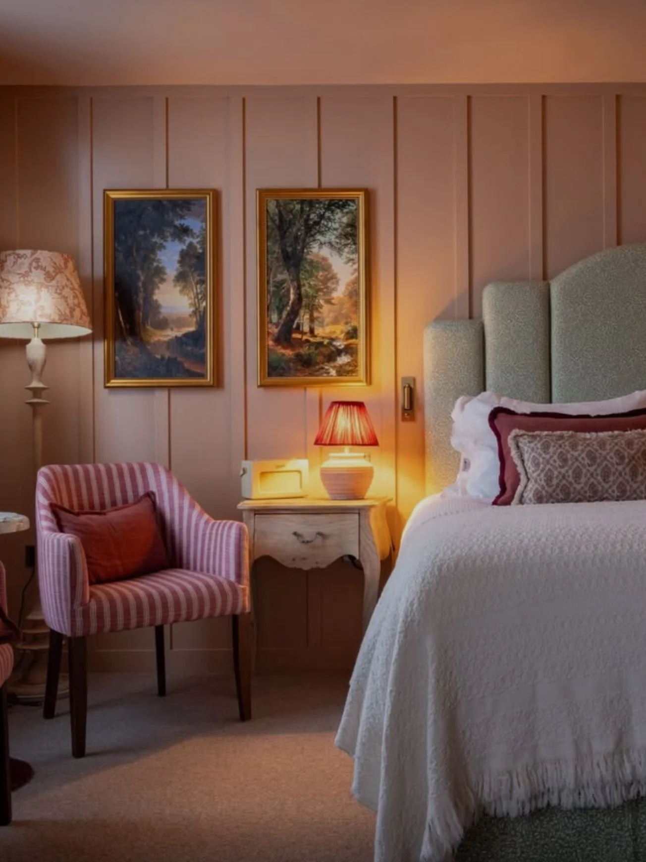 A cozy bedroom corner with a bed, a nightstand with a lit lamp, a pink striped armchair with a pillow, and two landscape paintings on the wall, illuminated by warm lighting.