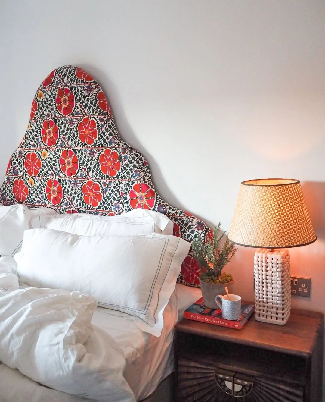 A cozy bedroom corner featuring a bed with white pillows and a crumpled white blanket, a wooden nightstand with a lamp, a small potted plant, a book titled 'The New Creative Home,' and a coffee mug, with a decorative fabric headboard behind the bed and blank white walls.