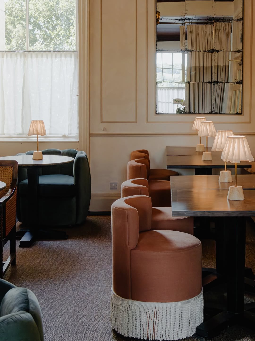 Interior of a cozy cafe or restaurant with tables, colorful upholstered chairs, and soft lamps, large windows with curtains, and a decorative mirror on the wall.