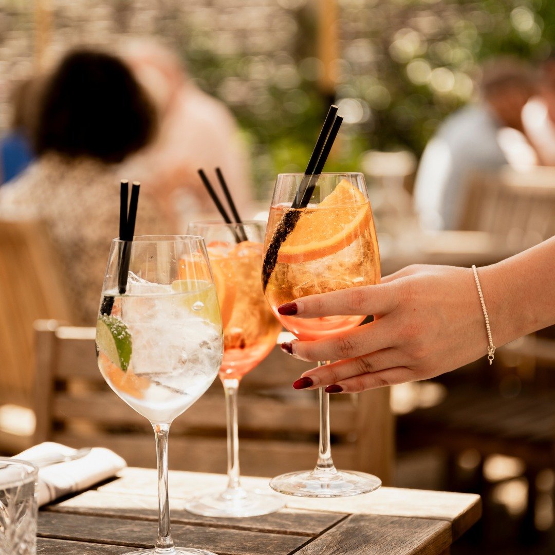Three cocktail glasses on a wooden table with drinks and straws, one glass is being held by a person with painted nails, in an outdoor setting with blurry background of people and trees.