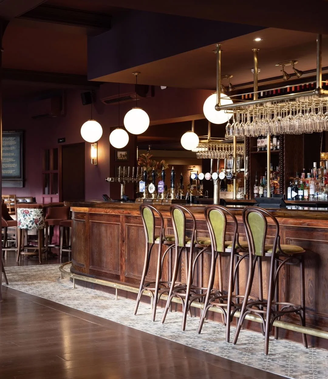 Empty bar with wooden counter, green upholstered bar stools, hanging glasses, liquor bottles, and round pendant lights in a dimly lit setting.