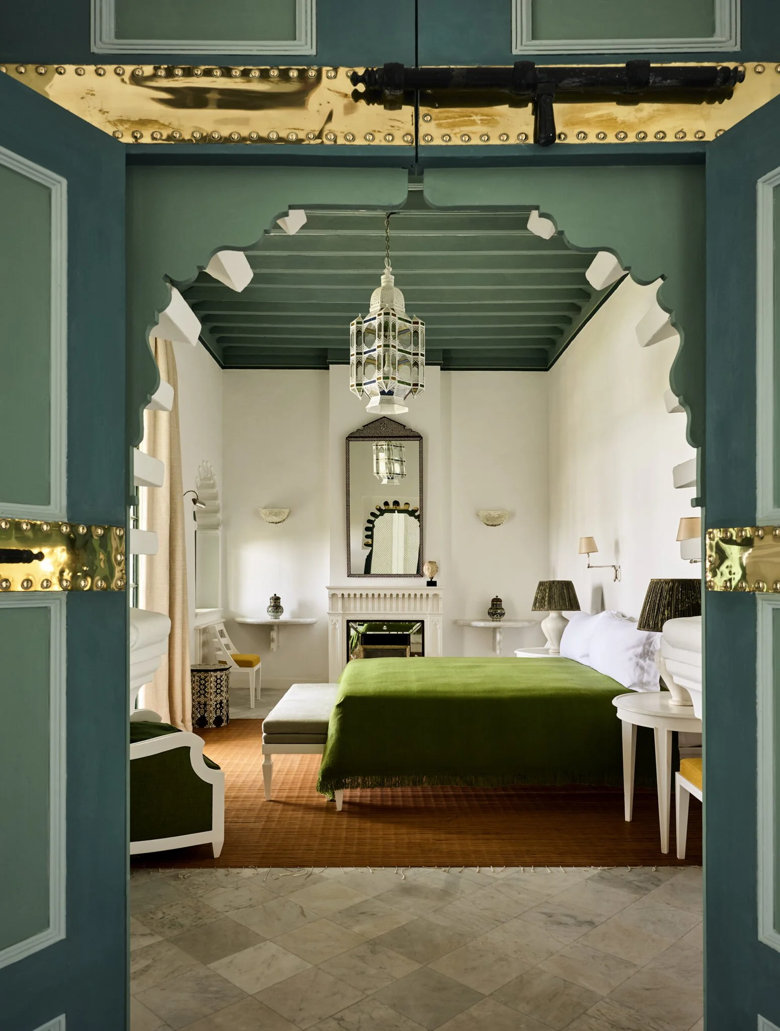 View through teal double doors into a bright, stylish bedroom with a green bedspread, white furniture, and Moroccan-style pendant lights, with a mirror and some decorative objects visible.