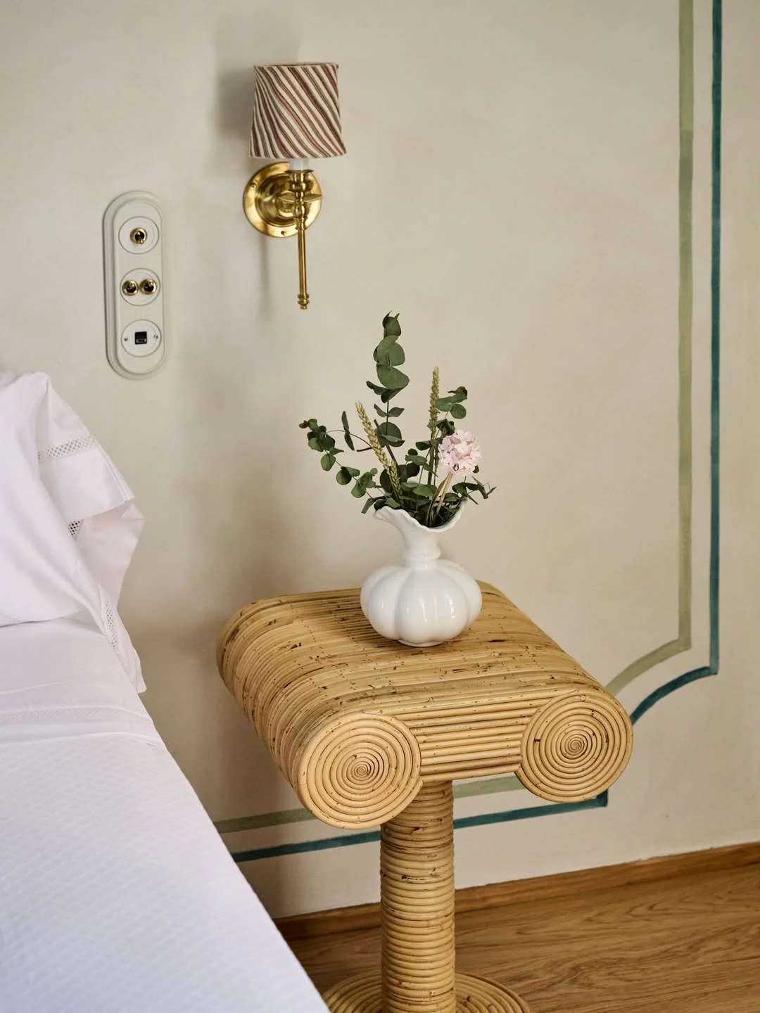 A white ceramic vase with pink flowers and green foliage on a rounded, wooden side table.