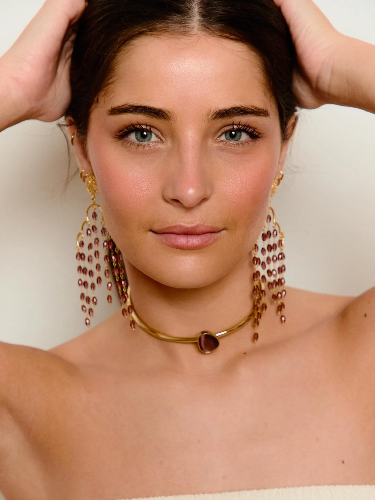 A close-up of a woman with short dark hair, wearing jewelry including gold earrings, a choker, and layered necklaces with a gemstone, and holding her head with both hands.