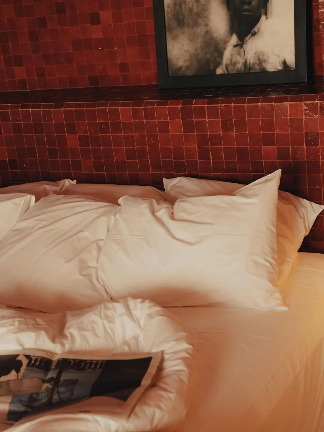 Unmade bed with white pillows and blankets in front of a red tiled headboard, with a black-and-white portrait photograph hanging above.