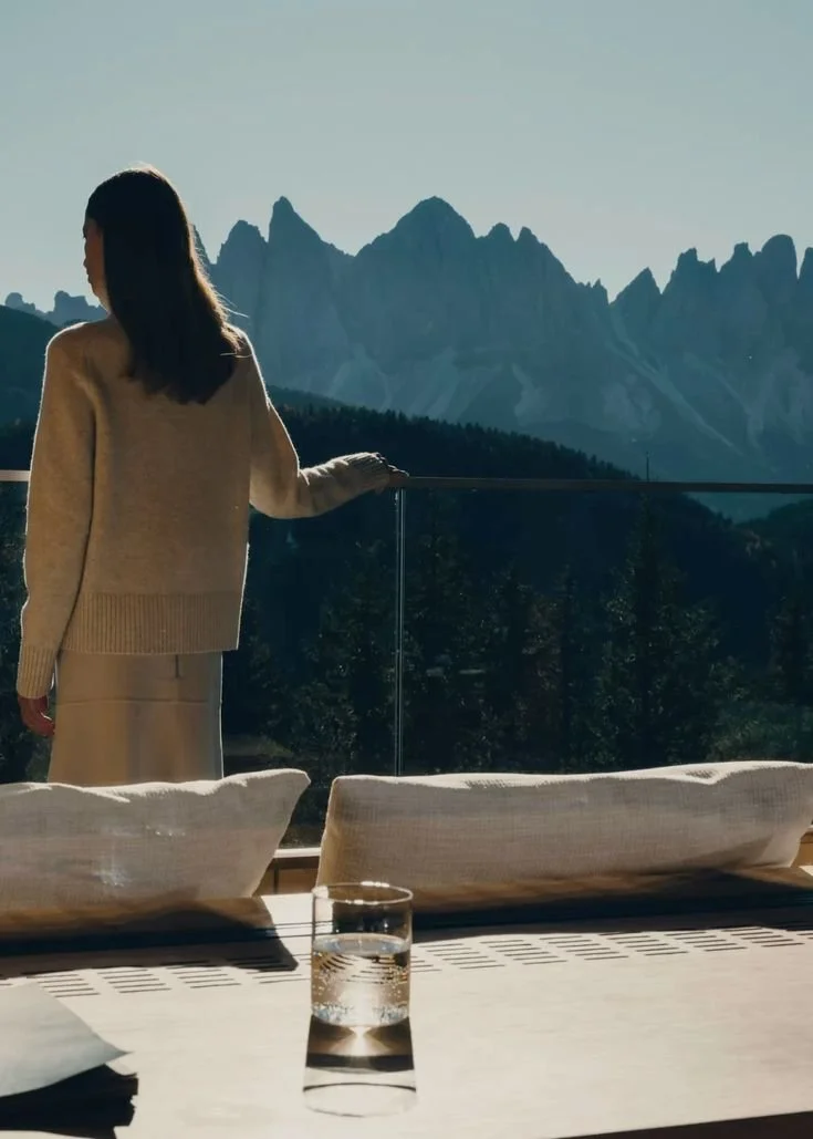 A woman standing on a balcony, gazing at mountain peaks in the distance, with a glass of water and cushions on a wooden table in the foreground.