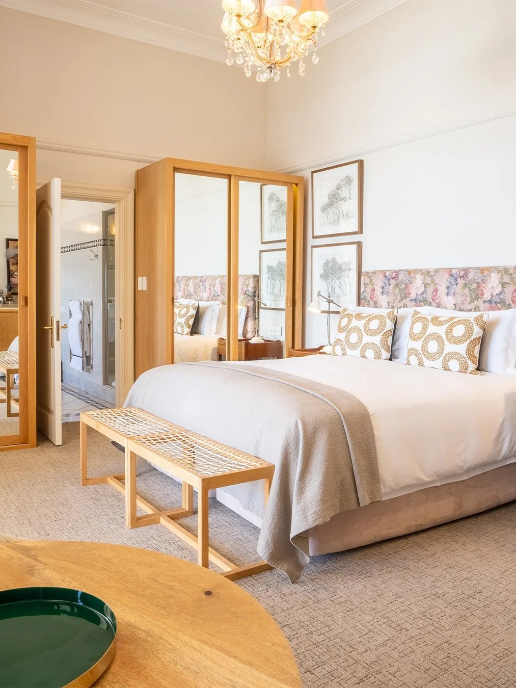 A cozy hotel bedroom with a large bed, decorated with patterned pillows and a floral headboard, wooden accents, a chandelier, and an attached bathroom visible through an open door.