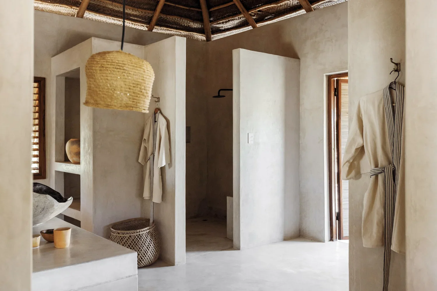 A minimalist bathroom with beige plaster walls and wooden accents, featuring several small shower stalls, hanging robes, wicker basket, and natural lighting from wooden window shutters.