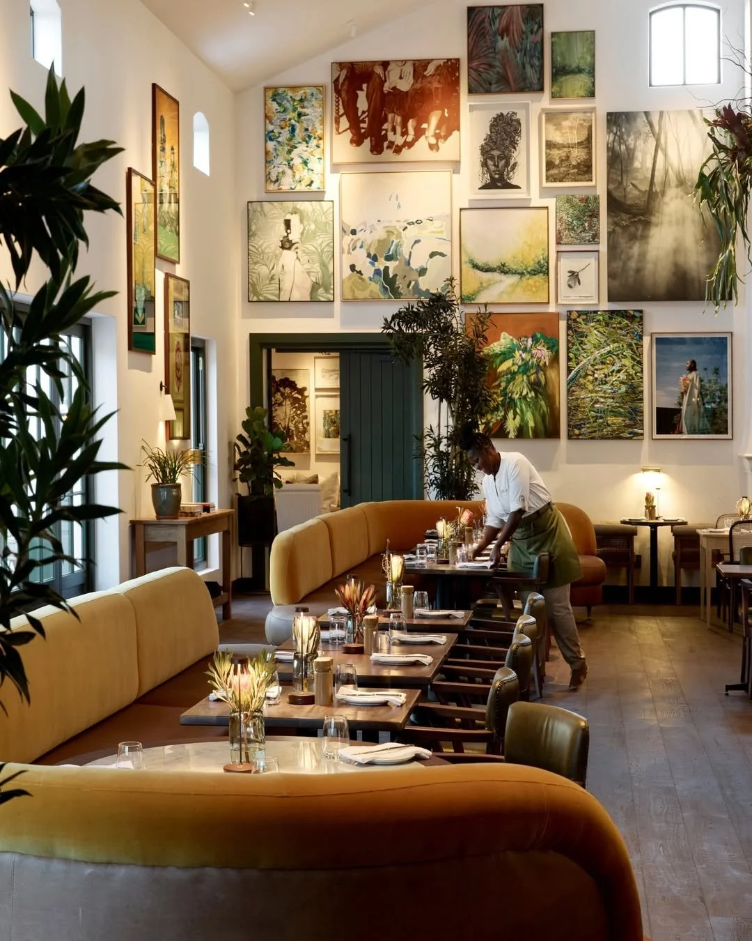 A cozy restaurant interior with a wall decorated with numerous framed artworks. A server is preparing tables, and the room has warm lighting and green plants.