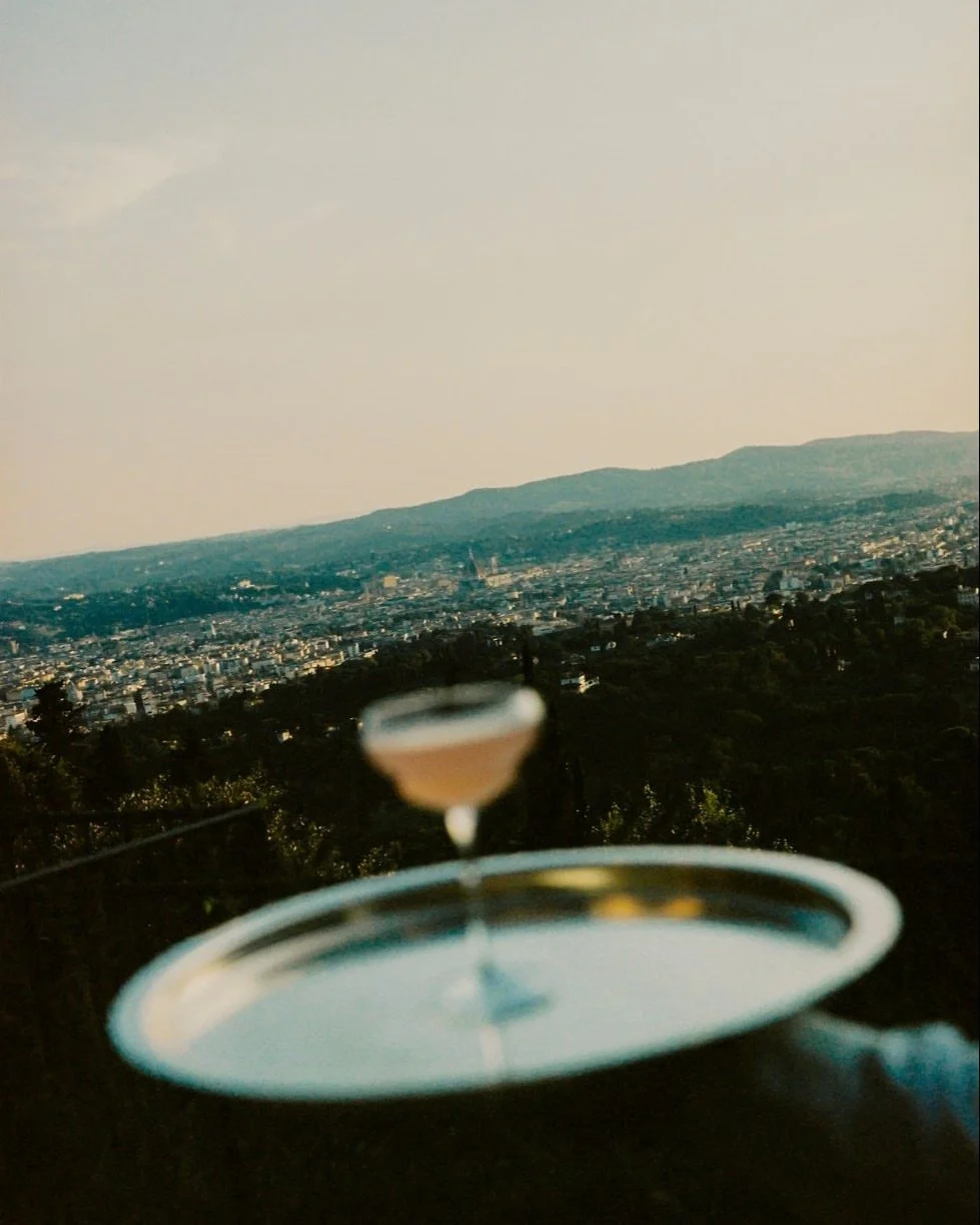 A blurred cocktail glass on a tray with a cityscape and mountains in the background during sunset.