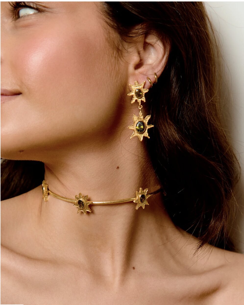 Close-up of a woman's face and neck showing gold jewelry with star-shaped designs and dark-colored stones.